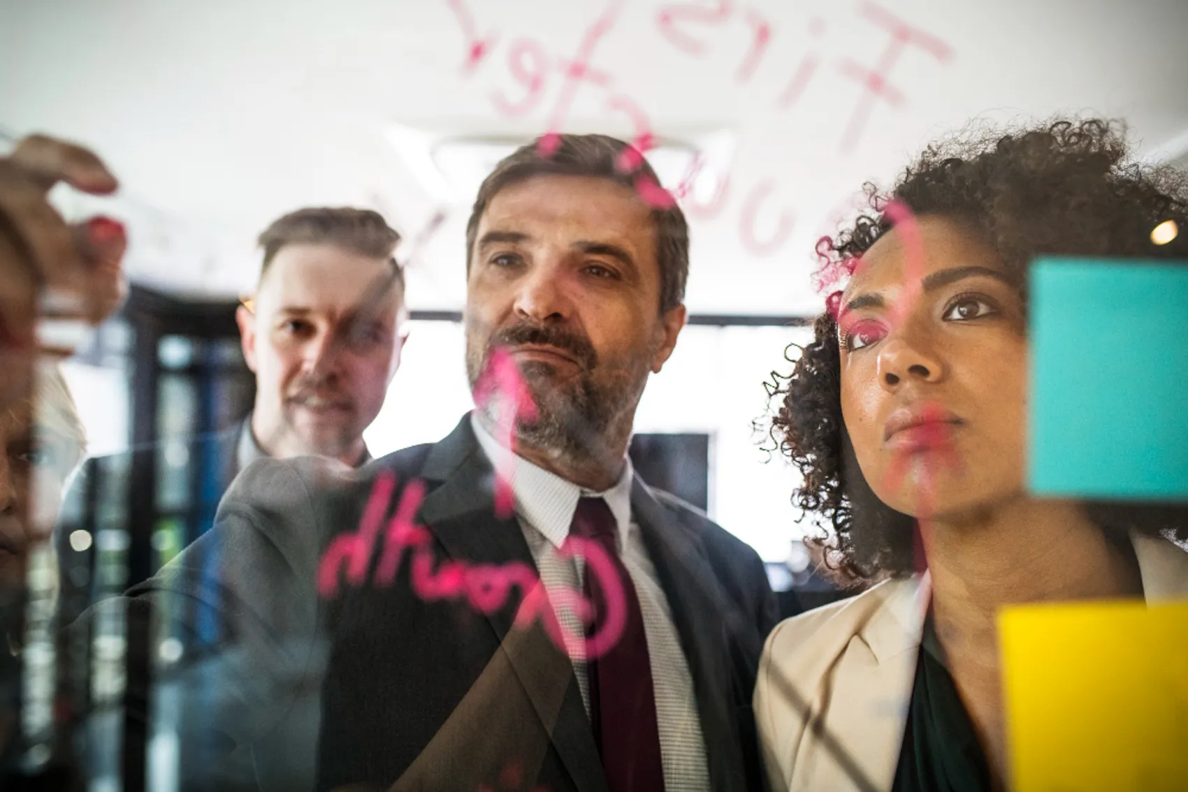 Três pessoas em ambiente corporativo, observando e escrevendo em vidro transparente com marcadores coloridos. No vidro, há palavras e símbolos escritos em rosa, incluindo a palavra 'Growth'. Também estão visíveis notas adesivas coloridas, como amarelo e azul, sugerindo uma sessão de brainstorming ou planeamento estratégico. Ao fundo, o espaço é iluminado por luz natural proveniente de janelas