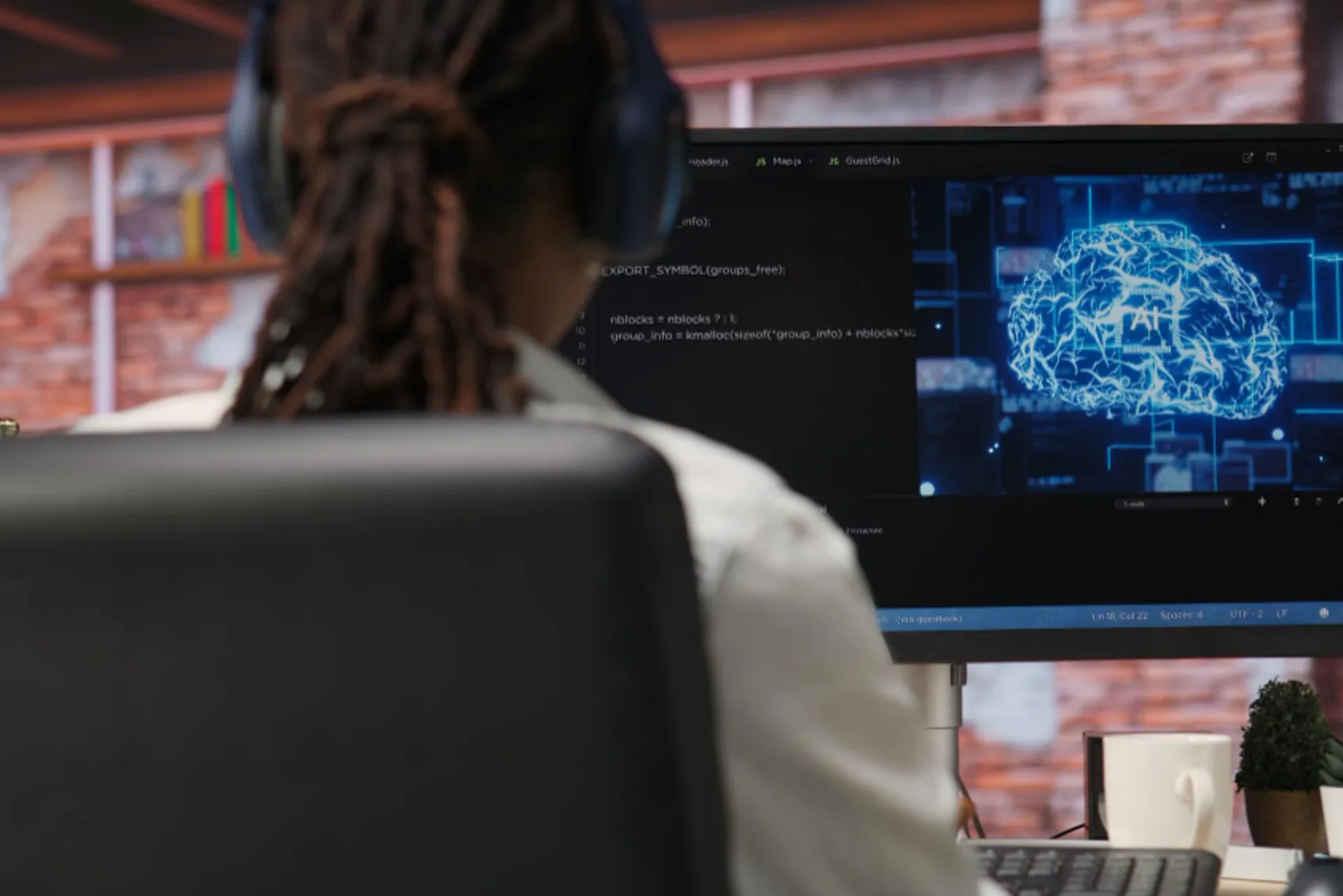 Pessoa sentada à secretária, usando auscultadores, a trabalhar num computador com monitor que exibe código e uma imagem digital de um cérebro estilizado em azul, representando inteligência artificial ou redes neurais. Sobre a mesa há um teclado, uma caneca branca e um pequeno vaso com planta. O fundo mostra uma parede com padrão de tijolos e prateleiras decorativas.